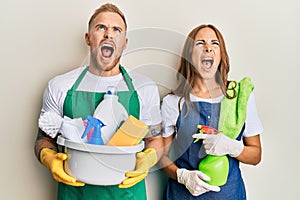 Young couple of girlfriend and boyfriend wearing apron holding products and cleaning spray angry and mad screaming frustrated and