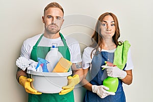 Young couple of girlfriend and boyfriend wearing apron holding products and cleaning spray puffing cheeks with funny face