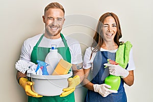 Young couple of girlfriend and boyfriend wearing apron holding products and cleaning spray winking looking at the camera with sexy