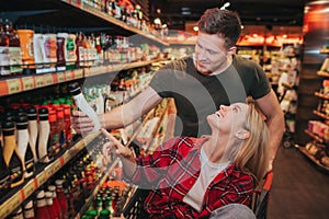 Young couple in grocery store. They pick sauce together. Young woman sit in trolley and look at man. Guy hold bottle