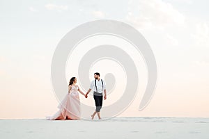 Young couple a guy in black breeches and a girl in a pink dress are walking along the white sand