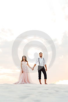 Young couple a guy in black breeches and a girl in a pink dress are walking along the white sand