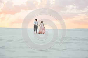 Young couple a guy in black breeches and a girl in a pink dress are walking along the white sand