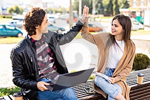 Young couple with hands up doing high five outdoor sitting with laptop on the bench. Concept of teamwork and fun together