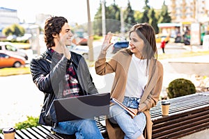 Young couple with hands up doing high five outdoor sitting with laptop on the bench. Concept of teamwork and fun together