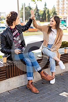 Young couple with hands up doing high five outdoor sitting with laptop on the bench. Concept of teamwork and fun together