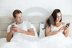 Young couple handsome husband and his beautiful wife just woke up in bed checking their smartphones early in the morning