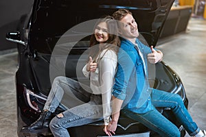 Young couple handsome man and beautiful woman sitting on open car bonnet both showing like signs in electric car dealership center