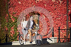 Young couple, handsome man and blond woman cycling tandem bike by building overgrown with red ivy.