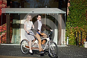 Young couple, handsome man and blond woman cycling tandem bike by building overgrown with red ivy.