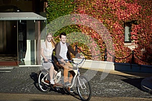 Young couple, handsome man and blond woman cycling tandem bike by building overgrown with red ivy.