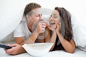 Young couple handsome man feeding beautiful woman popcorn with his hand watching movie in bed looking happy