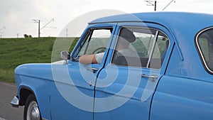 Young couple in hats driving at blue retro car after rain during summer travel. Unrecognizable pair moving in old