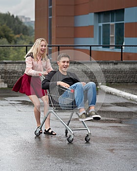 Young couple having fun on shopping trolley. Happy woman pushing shopping cart with her boyfriend inside