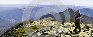 Young couple hike in Carpathian mountains. Man and woman standing on mountain top looking at beautiful landscape below. Wide panor