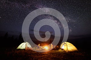 Young couple hikers resting near illuminated tent, camping in mountains at night under starry sky