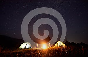 Young couple hikers resting near illuminated tent, camping in mountains at night under starry sky