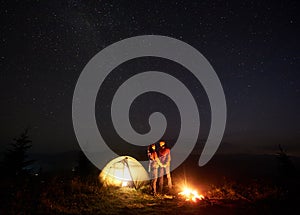 Young couple hikers resting near illuminated tent, camping in mountains at night under starry sky