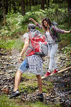 Young couple hiking in forest. Crossing a creek, boyfriend reaches out to help. Summertime, mountain landscape. Freedom,