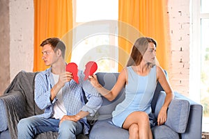 Young couple holding halves of broken heart on sofa at home. Relationship problems