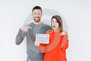 Young couple holding ovulation calendar over isolated background with surprise face pointing finger to himself