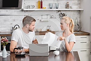 young couple holding papers and using laptop while sitting at kitchen table and looking photo