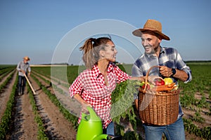 Young couple holding watering can and basket with vegetables and elderly farmer working in background
