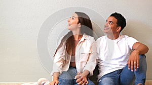 Young couple at home sitting on the floor looking up in to the air and smiling - concept of love and relationship with mortgage