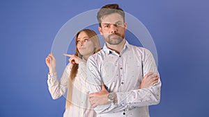 Young couple isolated on the blue background. Confidet man with crossed hands over the chests and woman making funny