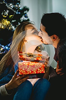Young couple kissing in front of Christmas tree, woman unpacking present