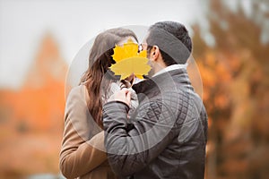 Young couple kissing, hiding faces with autumn yellow maple leave.