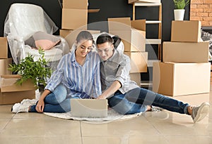 Young couple with laptop sitting on floor near boxes indoors. Moving into new house