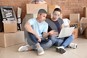 Young couple with laptop sitting on floor near boxes indoors. Moving into new house