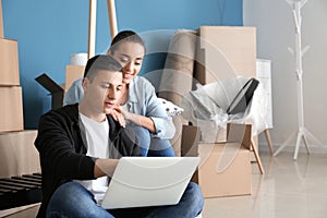 Young couple with laptop sitting on floor near boxes indoors. Moving into new house