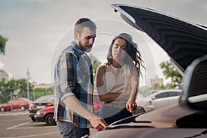 Young couple looking at the engine while standing near a broken car and an open hood outdoors. Car service, insurance