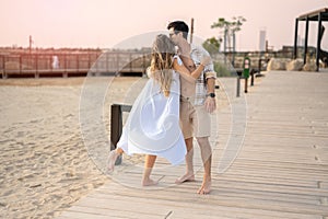 Young couple in love embracing while standing at beach walkway at sunset. Handsome man with kissing his girlfriend.