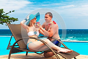 Young couple in love is enjoying cocktails at the poolside. Tropical sea in the background. Summer vacation.
