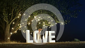 Young couple in love in evening dresses are dancing near love light letters.