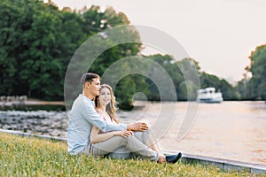 A young couple in love, a guy and a girl are sitting on the shore of the lake, happy and smiling. First date