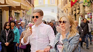 Young couple in love is having fun in the city center in front of old building. Brunette man feeds beautiful blonde