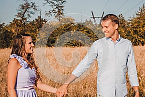 Young couple in love holding hands and smiling in hay field. Man leads woman forward. Outdoor portrait of beautiful romantic