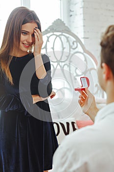 Young couple in love at home. Smiling man holding red box with marriage engagement ring got up on his knee and make proposal to