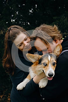 A young couple in love hugging with cute dog welsh corgi pembroke, exchanging loving glances. The dog is perched in the