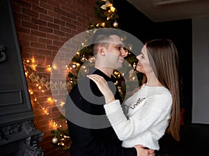 Young couple in love hugging by fireplace in front of Christmas tree, looking at each other, smiling. Handsome brunette man,