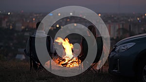 Young couple in love kissing sitting on chairs near bonfire at night enjoying warm romantic evening.