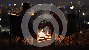 Young couple in love kissing sitting on chairs near bonfire at night enjoying warm romantic evening.