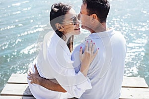 Young couple in love in light casual clothes is sitting on wooden bridge. Happy woman and man hug and laugh on background of sea