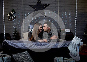 Young couple in love, lying on bed with christmas lights behind, embracing, kissing. Brunette man and woman, wearing grey winter