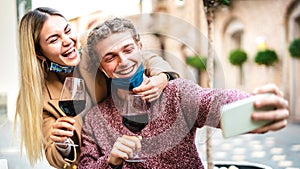 Young couple in love with open face mask taking selfie at outside wine bar - Happy millennial lovers enjoying lunch together