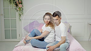 Young couple in love are sitting on bed in the bedroom and looking at the camera.